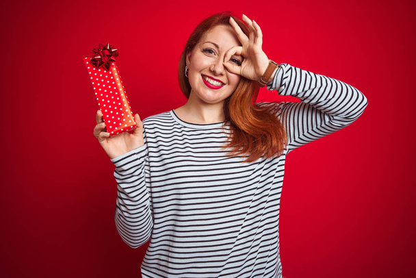 Nuori kaunis punapää nainen tilalla Ystävänpäivä lahja yli punainen eristetty tausta onnellinen kasvot hymyillen tekee ok merkki käsin silmään katselee sormien läpi
 - Valokuva, kuva