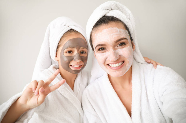 Mother and her little daughter in bathrobes and with facial masks against light background - Photo, Image