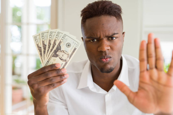 Afrikkalainen amerikkalainen mies jolla on kaksikymmentä dollaria seteleitä avoimella kädellä tekemässä stop-merkki vakavasti ja luottavainen ilme, puolustus ele
 - Valokuva, kuva