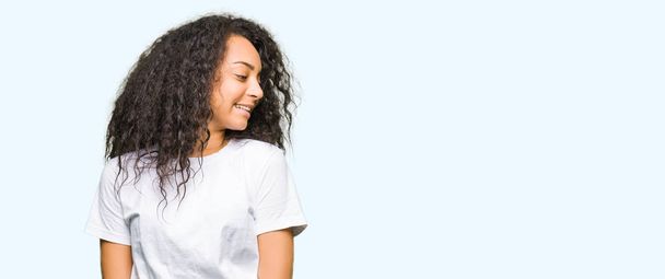 Nuori kaunis tyttö kihara tukka yllään rento valkoinen t-paita etsii sivuun hymyillen kasvot, luonnollinen ilme. Nauraen itsevarmana
. - Valokuva, kuva