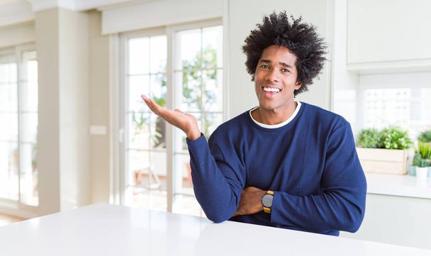Young african american man wearing casual sweater sitting at home smiling cheerful presenting and pointing with palm of hand looking at the camera. - Photo, Image