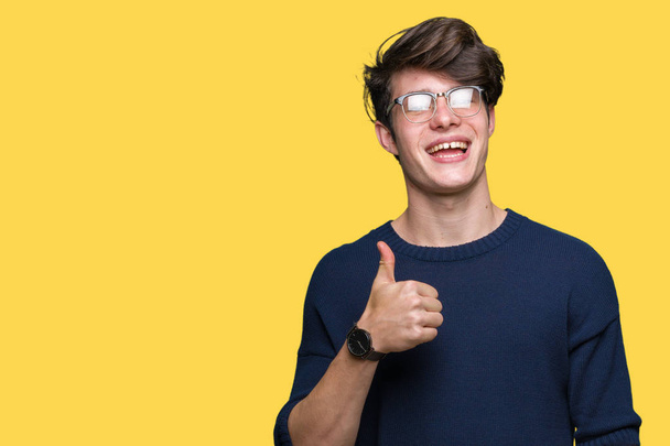 Mladý pohledný muž nosí brýle izolované pozadí dělá šťastné palec nahoru gesto rukou. Schvalování výraz při pohledu na fotoaparát ukazuje úspěch. - Fotografie, Obrázek