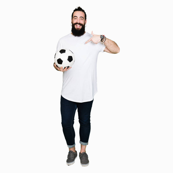 Giovane uomo con i capelli lunghi e orso in possesso di pallone da calcio con faccia a sorpresa puntando il dito verso se stesso
 - Foto, immagini