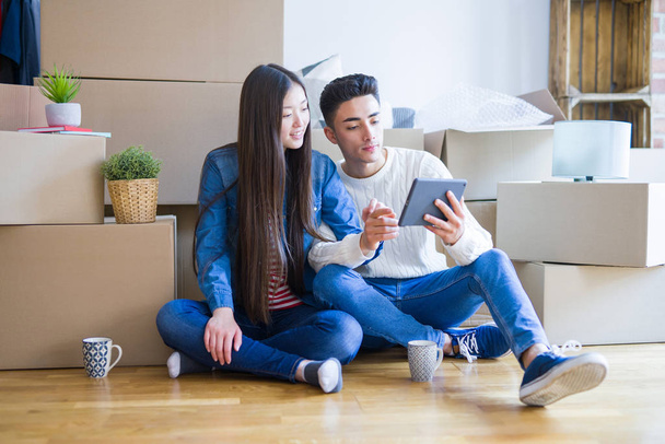 Young asian couple sitting on the floor of new apartment arround cardboard boxes, using touchpad tablet and smiling at new home - Fotó, kép