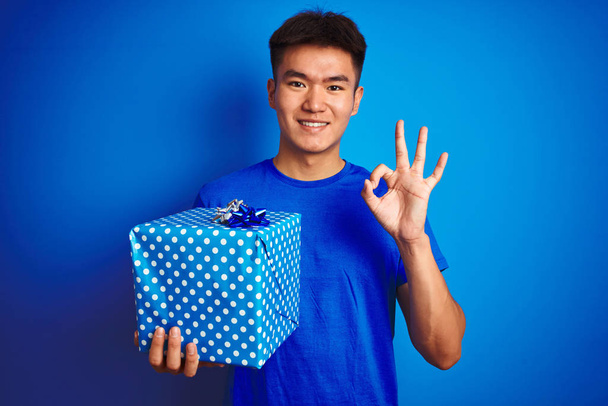 Young asian chinese man holding birthday gift standing over isolated blue background doing ok sign with fingers, excellent symbol - Foto, afbeelding