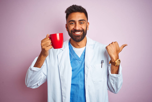 Young indian doctor man drinking cup of coffee standing over isolated pink background pointing and showing with thumb up to the side with happy face smiling - Photo, Image