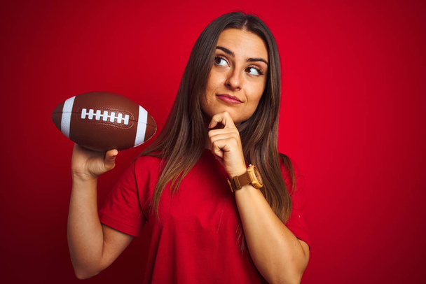 Mladá krásná sportovna, která drží fotbalový míč přes izolované červené pozadí seriózní tvář přemýšlími o otázce, velmi zmatené myšlence - Fotografie, Obrázek