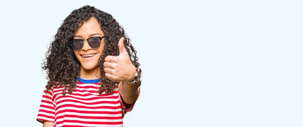 Young beautiful woman with curly hair wearing sunglasses doing happy thumbs up gesture with hand. Approving expression looking at the camera with showing success. - Photo, Image