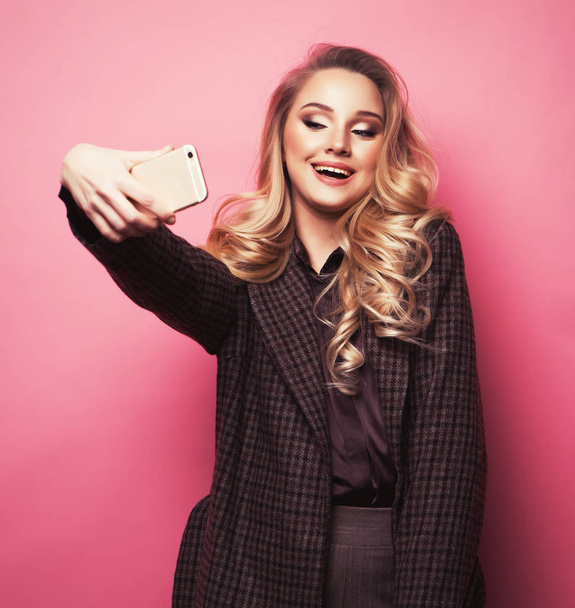 Blond pretty girl taking photo makes self portrait on smartphone over pink background - 写真・画像