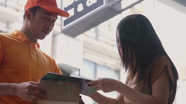 アジアの若い女性がクリップボードに署名を入れている間、段ボール箱を持つ笑顔の若い配達人. - 映像、動画