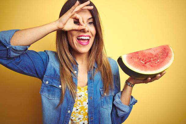 junge schöne Frau isst frische gesunde Wassermelonenscheibe auf gelbem Hintergrund mit glücklichem Gesicht lächelnd tut ok Zeichen mit Hand auf Auge durch Finger schauen - Foto, Bild