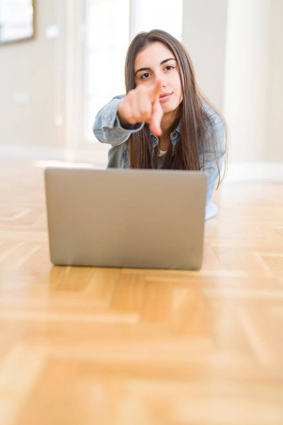 Kaunis nuori nainen makaa lattialla kannettavan tietokoneen avulla osoittaen sormella kameraa ja sinua, käsi merkki, positiivinen ja luottavainen ele edestä
 - Valokuva, kuva