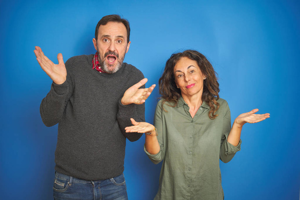Beautiful middle age couple together standing over isolated blue background clueless and confused expression with arms and hands raised. Doubt concept. - Photo, Image