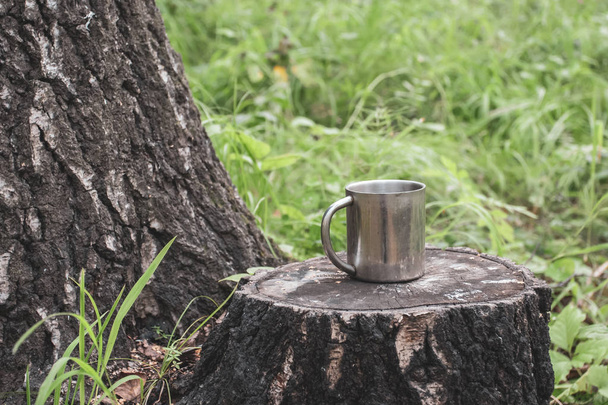 An iron tourist mug stands on a stump in a forest next to a thick tree against a background of blurred grass. Selective focus on the mug. The background is blurry. Place for text. - Foto, imagen