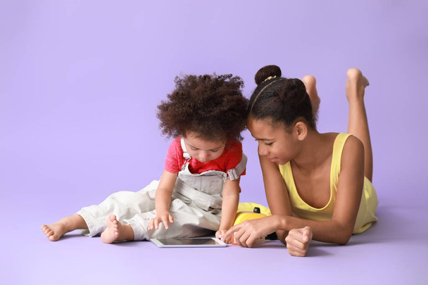 Cute African-American sisters with tablet computer watching cartoons on color background - Photo, Image