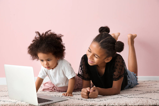 Cute African-American sisters with laptop watching cartoons near color wall - Foto, immagini