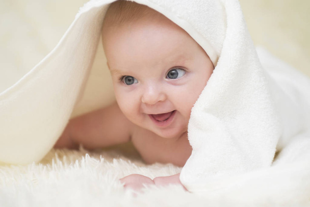 白いベッドの上にタオルで赤ちゃんを笑ってかわいいの肖像画。無邪気な赤ん坊は白いベッドの上をタオルで這う彼の頭の上に自宅で - 写真・画像