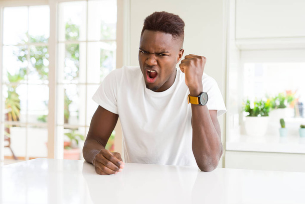 Hombre afroamericano guapo en la mesa blanca en casa enojado y loco levantando el puño frustrado y furioso mientras gritaba con ira. Rabia y concepto agresivo
. - Foto, imagen