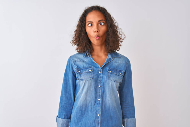 Young brazilian woman wearing denim shirt standing over isolated white background making fish face with lips, crazy and comical gesture. Funny expression. - Photo, image