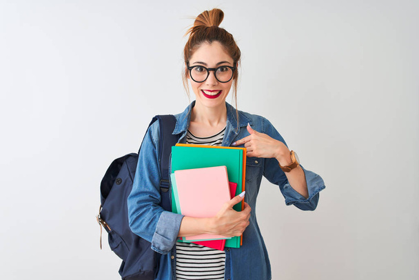 Estudante ruiva mulher usando mochila segurando livros sobre fundo branco isolado com rosto surpresa apontando dedo para si mesmo
 - Foto, Imagem
