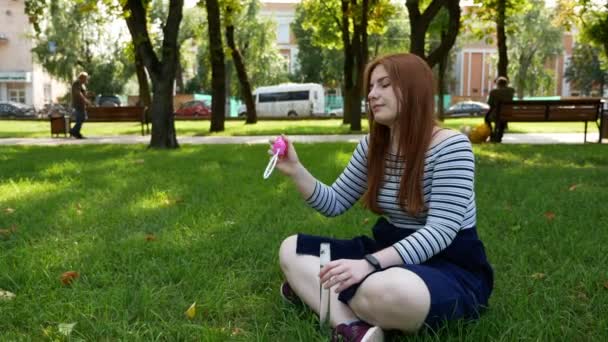 Κοκκινομάλλα κορίτσι χτυπήματα σαπούνι στο πάρκο χαμογελάει και γελάει το καλοκαίρι και την ευτυχία  - Πλάνα, βίντεο
