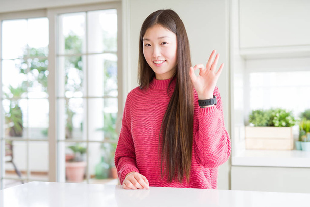 Piękna Azjatycka kobieta ubrana w różowy sweter na białym stole uśmiechnięty pozytywny robi OK znak z ręką i palcami. Pomyślne wyrażenie. - Zdjęcie, obraz