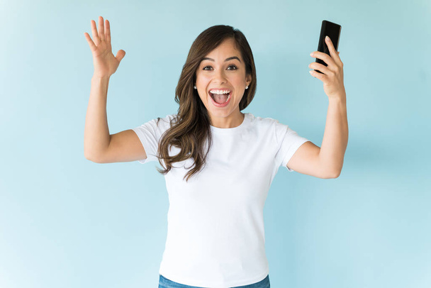 Cheerful excited woman holding smartphone while standing with arms raised against colored background - Fotografie, Obrázek