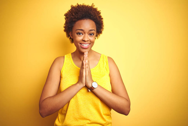 Beauitul femme afro-américaine portant t-shirt d'été sur fond jaune isolé priant avec les mains ensemble demandant pardon souriant confiant
. - Photo, image