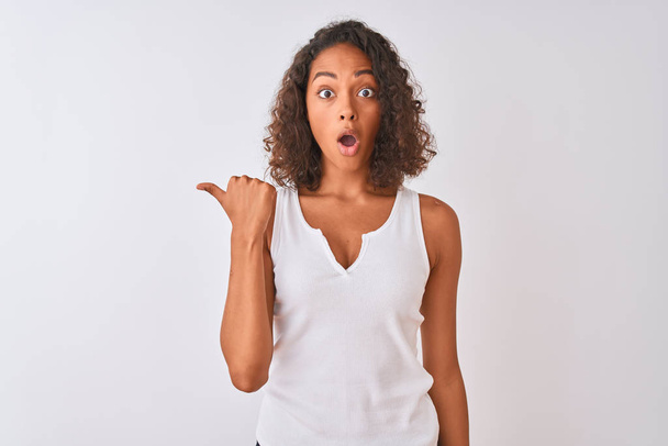 Jonge Braziliaanse vrouw dragen casual t-shirt staande over geïsoleerde witte achtergrond verrast wijzend met de hand vinger aan de zijkant, open mond verbaasd uitdrukking. - Foto, afbeelding