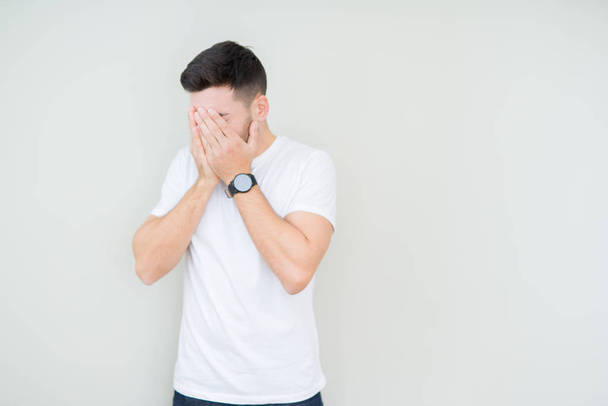 Jonge knappe man dragen casual wit t-shirt over geïsoleerde achtergrond met trieste uitdrukking waarin gezicht met handen terwijl het huilen. Depressie concept. - Foto, afbeelding
