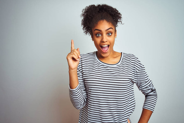 Mujer afroamericana vistiendo una camiseta de rayas navales de pie sobre un fondo blanco aislado señalando con el dedo hacia arriba con una idea exitosa. Salido y feliz. Número uno.
. - Foto, imagen