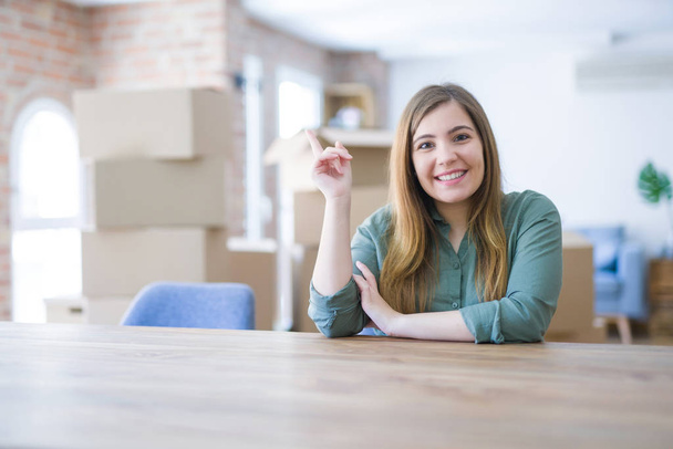 Mujer joven sentada en la mesa con cajas de cartón detrás de su mudanza a un nuevo hogar con una gran sonrisa en la cara, señalando con la mano y el dedo hacia el lado mirando a la cámara
. - Foto, Imagen
