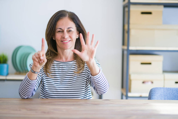 Femme âgée d'âge moyen assis à la table à la maison montrant et pointant vers le haut avec les doigts numéro six tout en souriant confiant et heureux
. - Photo, image