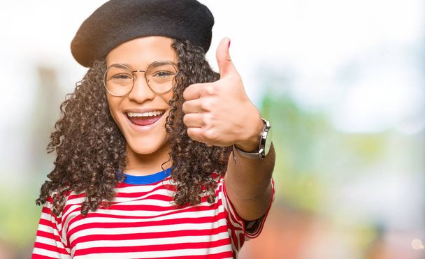 Young beautiful woman with curly hair wearing glasses and fashion beret doing happy thumbs up gesture with hand. Approving expression looking at the camera showing success. - Photo, Image