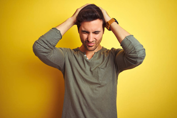Junger gutaussehender Mann in lässigem T-Shirt, der vor isoliertem gelben Hintergrund steht und unter Kopfschmerzen leidet, die verzweifelt und gestresst sind, weil Schmerzen und Migräne. Hände auf dem Kopf. - Foto, Bild