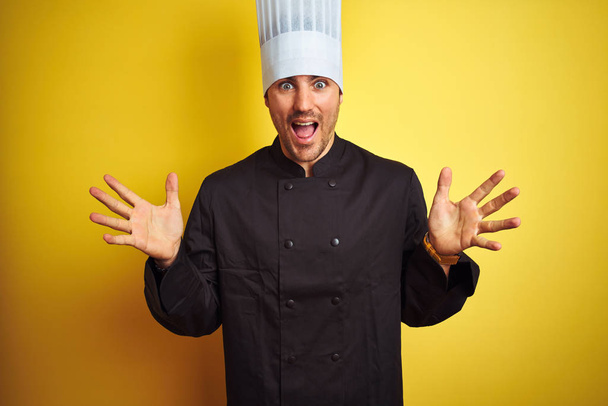 Young chef man wearing uniform and hat standing over isolated yellow background celebrating crazy and amazed for success with arms raised and open eyes screaming excited. Winner concept - Photo, Image