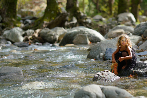 3歳の少女はユービーアのディルフィ山の美しい自然の中で川の近くに古い人形で遊んで - 写真・画像