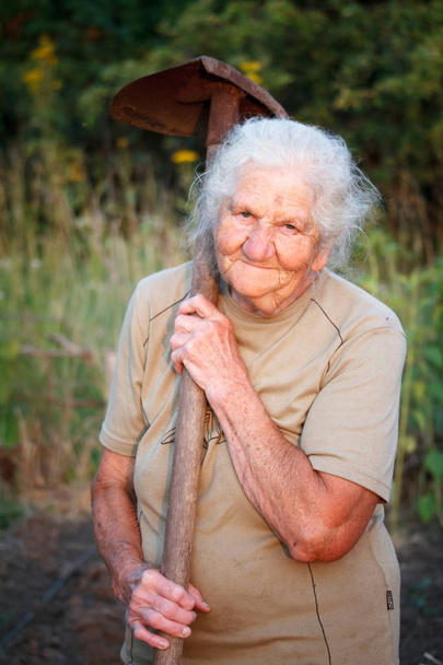 Close-up retrato de uma mulher idosa com cabelos grisalhos sorrindo e olhando para a câmera, segurando uma pá enferrujada em suas mãos, rosto em rugas profundas, foco seletivo
 - Foto, Imagem