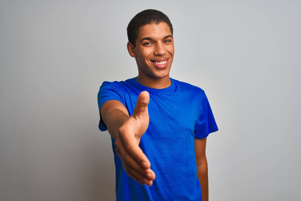 Mladý pohledný arabský muž, oblečený do modrého trička, stojící nad ojedinělými bílými pozadí, usměvavý, nabízející stisk ruky jako pozdrav a přívětivý. Úspěšný obchod. - Fotografie, Obrázek
