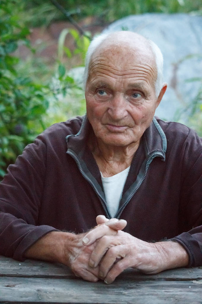 Retrato de cerca de un hombre muy anciano con los brazos cruzados, sentado en una mesa sobre un fondo borroso de árboles verdes, enfoque selectivo
 - Foto, Imagen
