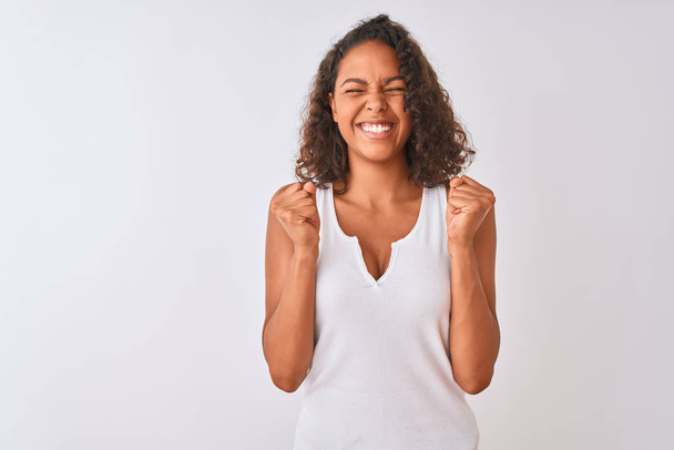 Nuori brasilialainen nainen päällään rento t-paita seisoo yli eristetty valkoinen tausta innoissaan menestystä kädet nostetaan ja silmät kiinni juhlii voittoa hymyillen. Voittajan käsite
. - Valokuva, kuva