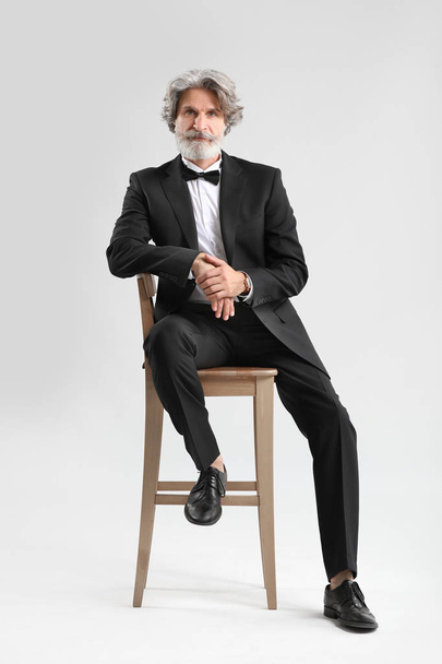 Modieuze Senior man in stijlvolle pak zittend op stoel tegen lichte achtergrond - Foto, afbeelding