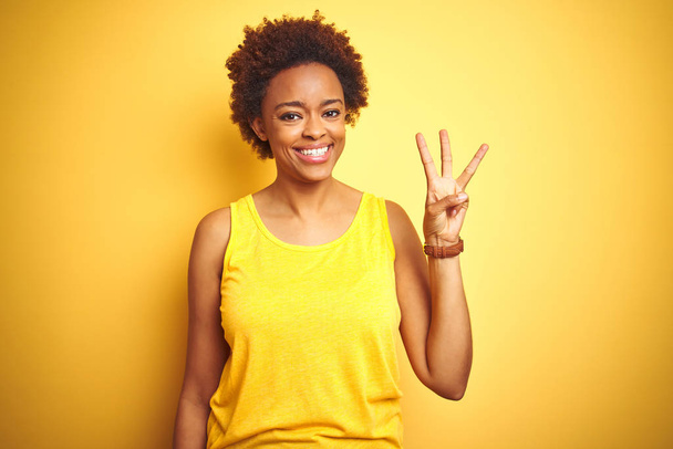 Африканская женщина Бойтул носит летнюю футболку на изолированном желтом фоне и показывает пальцами номер три, улыбаясь уверенно и счастливо
. - Фото, изображение