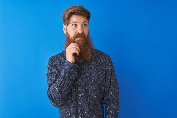 Jonge Redhead Ierse man dragen Floral zomer shirt staande over geïsoleerde blauwe achtergrond met de hand op kin denken over vraag, pensieve uitdrukking. Glimlachend met doordachte gezicht. Doubt concept. - Foto, afbeelding