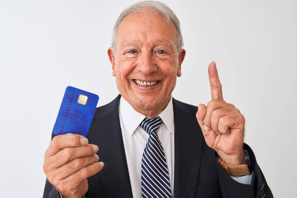 Senior grey-haired businessman holding credit card over isolated white background surprised with an idea or question pointing finger with happy face, number one - Zdjęcie, obraz