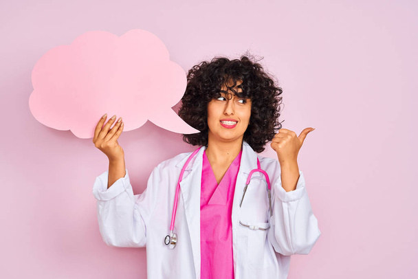 Mujer árabe joven con el pelo rizado sosteniendo la burbuja del habla de la nube sobre el fondo rosa aislado señalando y mostrando con el pulgar hacia arriba al lado con la cara feliz sonriendo
 - Foto, imagen
