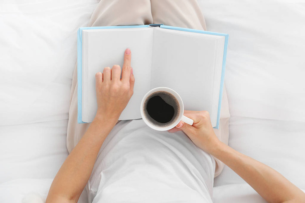schöne junge Frau trinkt Kaffee, während sie Buch im Schlafzimmer liest, Draufsicht - Foto, Bild