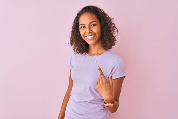Mujer brasileña joven vistiendo camiseta de pie sobre fondo rosa aislado Beckoning venir aquí gesto con la mano invitando a la bienvenida feliz y sonriente
 - Foto, Imagen