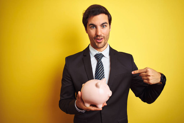 Jonge knappe zakenman Holding Piggy Bank over geïsoleerde gele achtergrond met verrassing gezicht wijzende vinger naar zichzelf - Foto, afbeelding