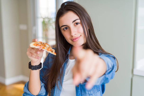 Mulher bonita comendo uma fatia de pizza saborosa apontando com o dedo para a câmera e para você, sinal de mão, gesto positivo e confiante da frente
 - Foto, Imagem